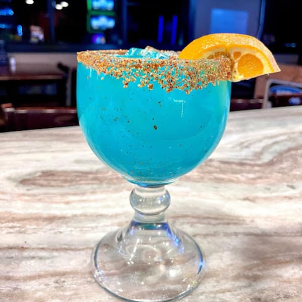 A vibrant blue Mayan Palace margarita with a spiced rim and orange slice, on a marble counter.