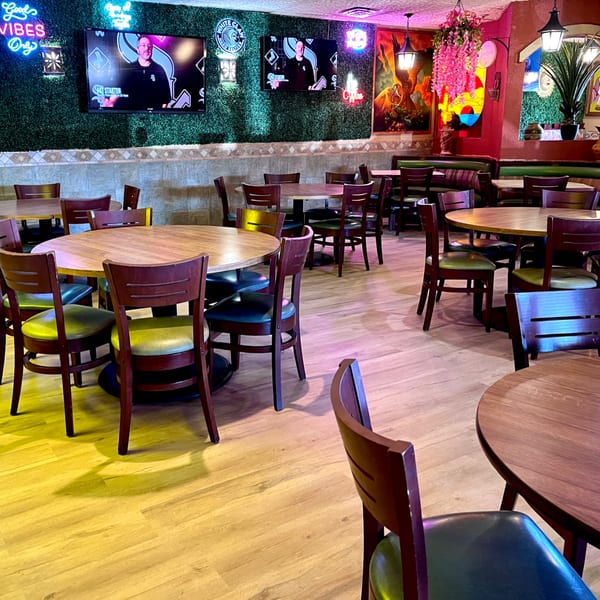 Interior of Mayan Palace with round wooden tables, green chairs, and neon signs on a green wall.