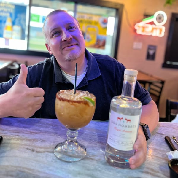 Man giving a thumbs up, holding a Dos Hombres mezcal bottle next to a Mayan Palace margarita.