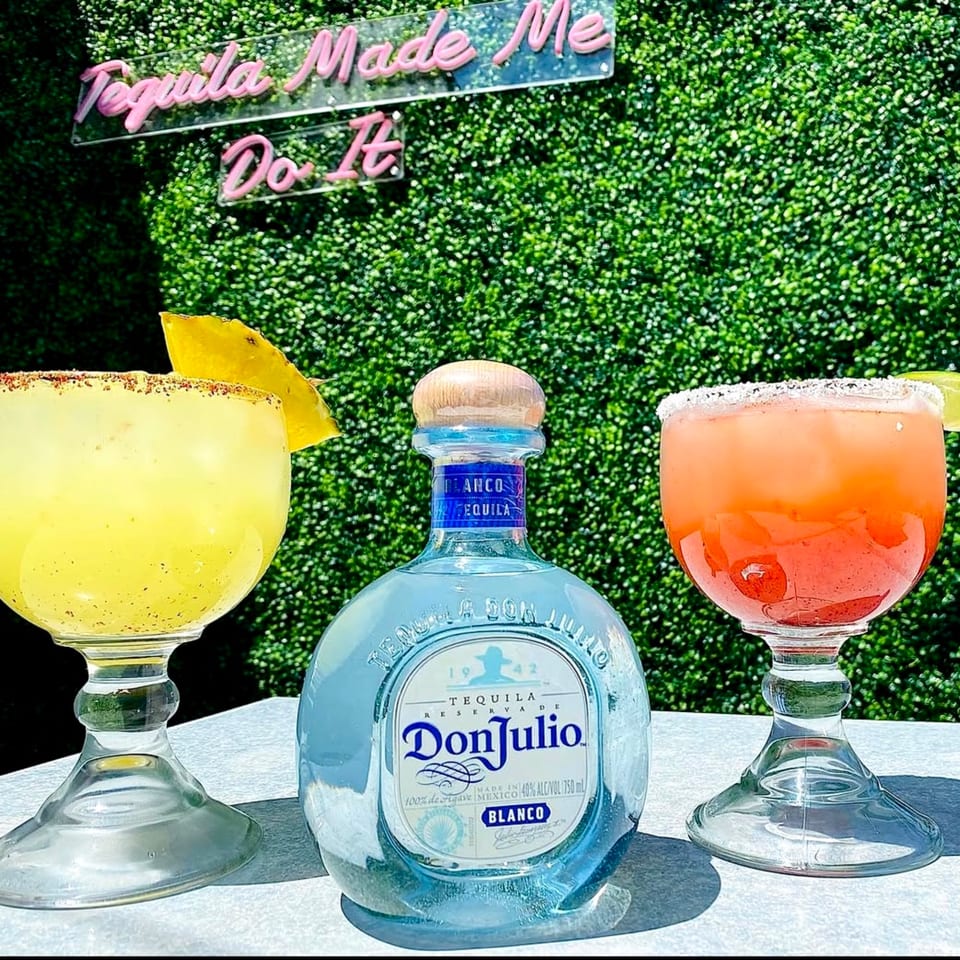 Don Julio tequila bottle between yellow and orange margaritas with a "Tequila Made Me Do It" sign.