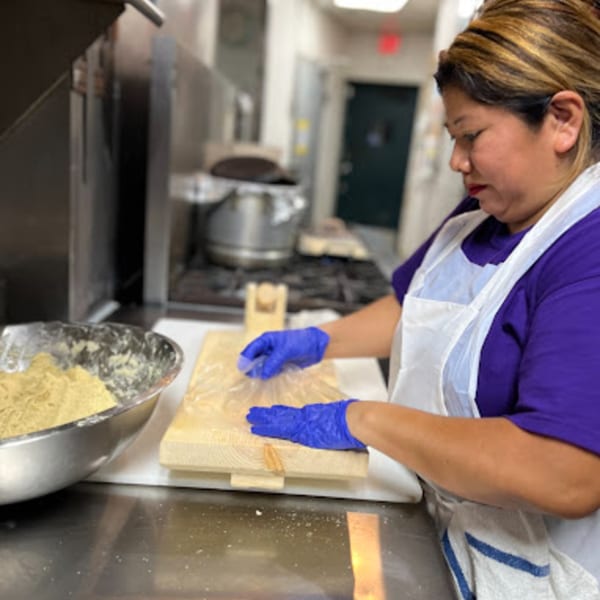 A Mayan Palace chef in a purple shirt and white apron presses masa dough on a wooden tortilla press.
