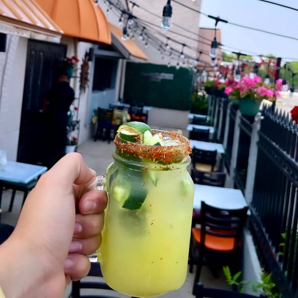 Hand holding a Mayan Palace cucumber margarita with chili-lime rim on an outdoor patio.
