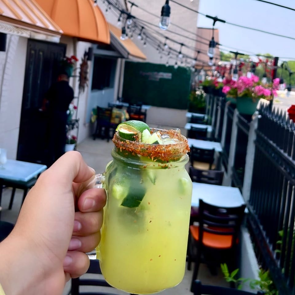 Hand holding a Mayan Palace cucumber margarita with chili-lime rim on an outdoor patio.