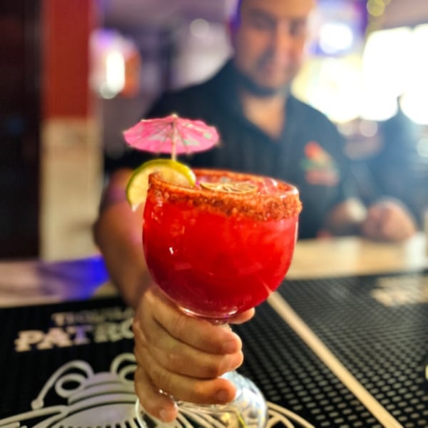 A hand holds out a vibrant red Mayan Palace margarita with a lime, umbrella, and chili rim.