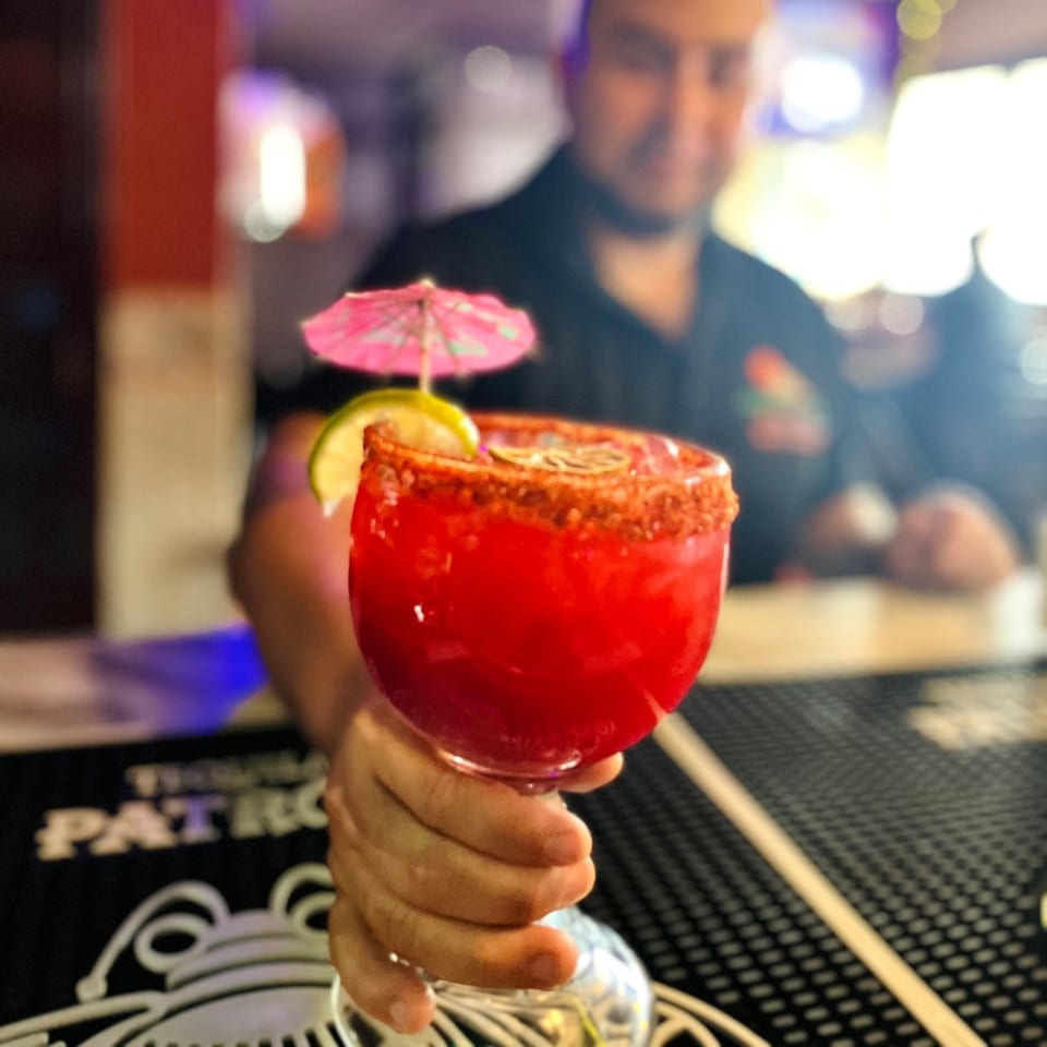 A hand holds out a vibrant red Mayan Palace margarita with a lime, umbrella, and chili rim.