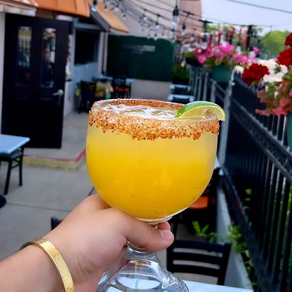 A hand holds a Mayan Palace margarita with a chili-lime rim and lime wedge on a patio.